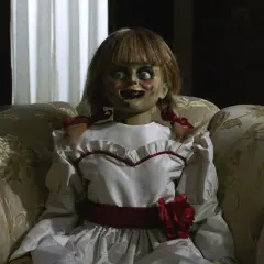 Den läskiga riktiga Annabelle dockan sitter i en blommig soffa, med sina stora ögon och röda rosett.