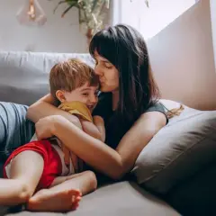 Mama całuje synka w czoło, przytulając go na kanapie. Dziecko śmieje się radośnie. Może dziś nie posyłam dziecka do przedszkola.
