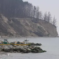 Klif Orłowski z łodziami rybackimi na kamienistym brzegu. Na zboczu klifu widać nagie drzewa i spacerujących ludzi.