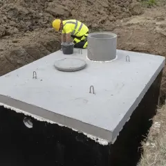Budowa betonowego zbiornika na deszczówkę. Pracownik w żółtym kasku i kamizelce odblaskowej nadzoruje montaż.