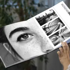 Osoba przegląda album "Mgnienie Oka", gdzie obok zbliżenia oka kobiety znajdują się czarno-białe fotografie miejskich krajobrazów i ruchu.