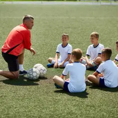 Trener w czerwonej bluzie prowadzi szkolenia dla trenerów, instruując młodych piłkarzy siedzących na boisku.