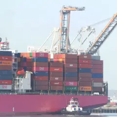Ogromny kontenerowiec z napisem UASC, załadowany po brzegi kolorowymi kontenerami, manewruje przy pomocy holownika.
