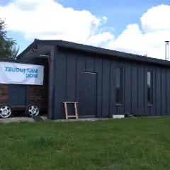 Mobilny garaż bez pozwolenia, z banerem "Zbuduj sam dom", stoi na trawie.