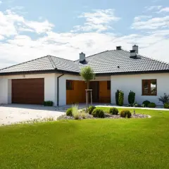 Nowoczesna budowa domu parterowego z garażem, białymi ścianami i drewnianą bramą, otoczona zielonym trawnikiem i zadbanym ogrodem.