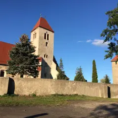 Kamienna wieża kościelna z czerwonym dachem w Nowogród Bobrzański, otoczona murem i zielenią.