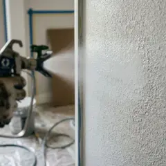 Mano guantata con pistola a spruzzo per imbiancare a spruzzo una parete ruvida.