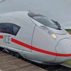 Biały pociąg Siemens Velaro ICE z czerwonym pasem stoi na drewnianym peronie. W tle pole i zachmurzone niebo.