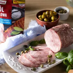 Pyszna szynka z szynkowara, pokrojona w plastry, podana z oliwkami, kaparami i świeżą bazylią. Obok opakowanie soli peklowej Prymat.