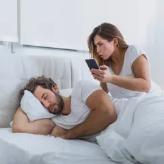 Donna preoccupata controlla il telefono del partner addormentato, un classico esempio di psicologia della sfiducia.