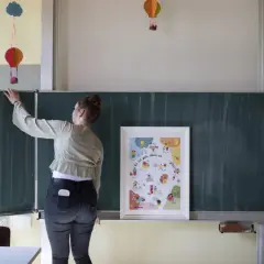 Lehrerin vor Tafel mit Poster "Es ist gut, dass es mich gibt!". Bunte Ballons hängen von der Decke. Ein Beispiel für heilende Klassenzimmer.