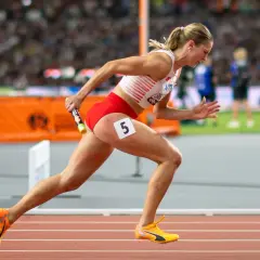 Zawodniczka biegnie z pałeczką w sztafecie 4x100 kobiet. Numer 5 na udzie.