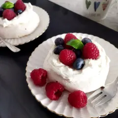 Dwa torty bezowe pavlova z bitą śmietaną, malinami i borówkami na talerzykach.