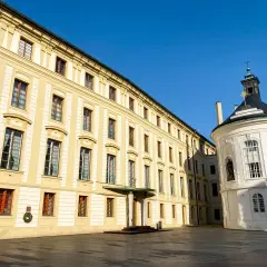 Hradczany zamek w Pradze. Długi, żółty budynek z wieloma oknami i biała, okrągła kaplica pod błękitnym niebem.