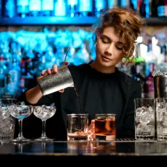 Kto robi drinki? Młoda barmanka z kręconymi włosami przelewa płyn z shakera do szklanki z lodem.