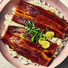 Two glazed unadon fillets rest on a bed of white rice, garnished with green onions and pickled ginger.