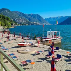 Plaże nad jeziorem Como, z rzędami leżaków i parasoli, czekają na turystów. W tle góry i błękitne niebo.