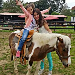 Niña feliz en un poni, disfrutando de una ruta a caballo en Cantabria. Madre sonriente la acompaña.