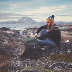 Mężczyzna w czapce i swetrze z norweskim wzorem siedzi na skale, podziwiając widok na kolorowe domy i góry. To jego wycieczka na Grenlandię.