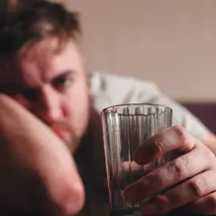 Mężczyzna z kacem trzyma pustą szklankę, opierając głowę na ręce.