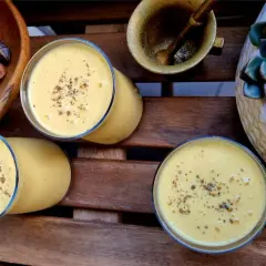 Trzy szklanki kremowego mango lassi, posypane przyprawami, obok miski z suszonymi owocami i sukulentu.