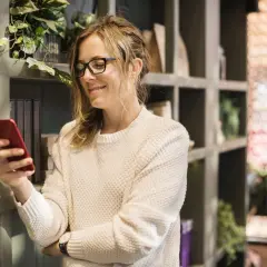 Kobieta w okularach uśmiecha się, trzymając czerwony telefon. Chyba prokrastynuję, zamiast czytać książki z półki.