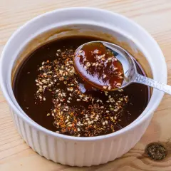 A spoon scoops rich, dark okonomiyaki sauce from a white bowl, sprinkled with sesame seeds and chili flakes. This is how to make okonomiyaki sauce.