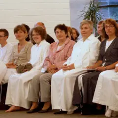 Frauen in weißen Gewändern sitzen nebeneinander. Die Kleidung könnte auf eine religiöse Gemeinschaft hindeuten, wo Baptisten Regeln für Frauen haben.