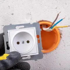 Ręka w rękawicy przygotowuje podłączenie gniazdka. Widać przewody wychodzące z puszki instalacyjnej.