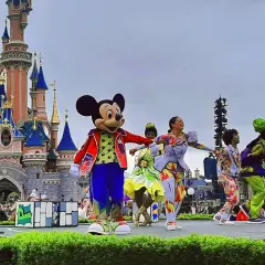 Myszka Miki i Goofy na scenie przed zamkiem w Disneyland Paris, otoczeni tancerzami w kolorowych strojach. Niezapomniane atrakcje Disneyland Paris!