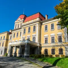 Żółty pałac z czerwonym dachem, otoczony zielenią i jesiennymi drzewami. W oddali widać kończyce wielkie.