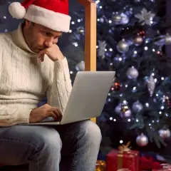 Mężczyzna w czapce Mikołaja siedzi przy choince, pracując na laptopie. Widać prezenty pod choinką. To smutne **święta**.
