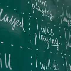Scrittura a mano su lavagna verde: "I played", "I was playing", "I have played", "I will play". Esercizi di grammatica inglese, con focus sui tempi verbali.