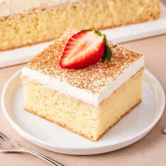 Ein Stück saftiger Tres Leches Kuchen, garniert mit einer Erdbeerhälfte und Zimt, auf einem Teller mit einer Gabel daneben.