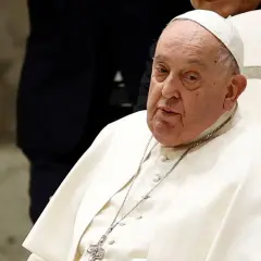 Papież Franciszek, siedzący na białym krześle, w białej sutannie i piusce. Jego wzrost jest typowy dla mężczyzny w jego wieku.