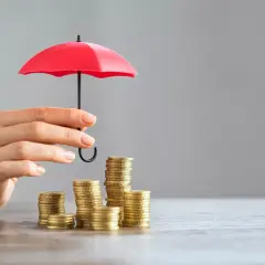 Une main protège des piles de pièces avec un petit parapluie rouge, symbolisant l'assurance PTZ.