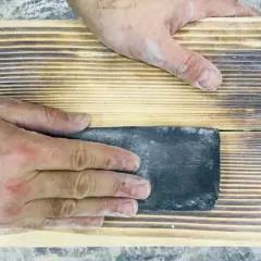 Mani che levigano un pezzo di legno con carta vetrata per sverniciare legno, rimuovendo la vernice vecchia.