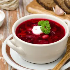 Pyszny botwinka przepis na rozgrzewającą zupę z buraków, podana z kleksem śmietany i natką pietruszki. Obok kromki chleba i czosnek.