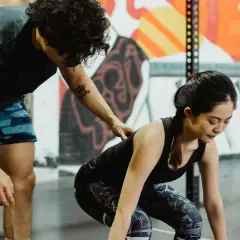 Entrenador ayuda a mujer a levantar pesas en gimnasio con mural colorido de fondo.