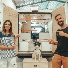 Pareja sonriente y su perro posan junto a una furgoneta camper en proceso de construcción, ¡las mejores furgonetas camper calidad precio!