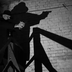 A man in a fedora and trench coat aims a pistol in this dramatic film noir photography. His shadow looms large against a brick wall.