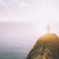 Ein Mensch steht auf einer Klippe am Meer, die Sonne scheint hell. Ein Symbol für das Durchhalten in schweren Zeiten, wenn man dem Horizont entgegenblickt.