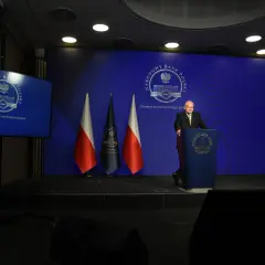 Prezes NBP Adam Glapiński przemawia na tle flag i ekranu z logo banku. Wiron 1m.