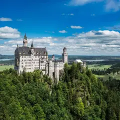 El castillo del rey loco, Neuschwanstein, se alza majestuoso sobre un risco verde, con torres que acarician las nubes y un paisaje de ensueño.