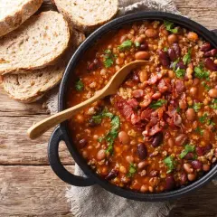 Pyszna fasolka po bretońsku w garnku, posypana boczkiem i natką pietruszki, obok kromki chleba.