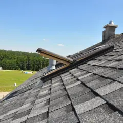 Dach mit Bitumenschindeln abdichten: Ein Dachfenster und ein Schornstein sind fachmännisch in die Schindeln integriert, um das Haus vor Regen zu schützen.