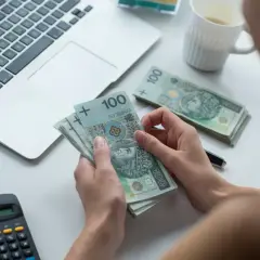 Kobieta liczy pieniądze, sprawdzając, czy kwota wolna od zajęcia jest wystarczająca. Obok leży laptop, okulary i kalkulator.