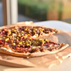 Pyszna pizza z kukurydzą, salami i oregano, podana na papierze do pieczenia, w słonecznym świetle.