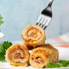 Roladki drobiowe z serem, zawinięte z szynką, prezentują się apetycznie na białym talerzu. Jedna z nich jest nabita na widelec.
