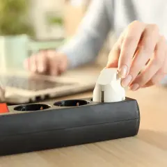 Dłoń podłącza wtyczkę do listwy zasilającej. Obok leży laptop, sugerując pracę lub naukę. Optymalizacja kosztów energii, np. w taryfie c11, jest ważna.